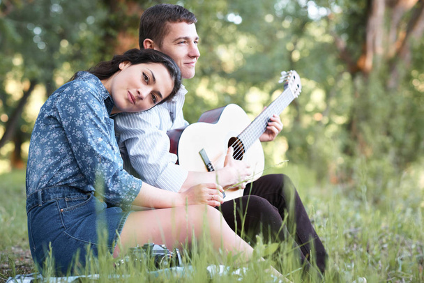 young couple sitting in the forest and playing guitar, summer nature, bright sunlight, shadows and green leaves, romantic feelings - Photo, Image