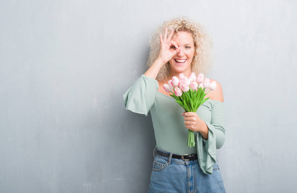 Young blonde woman over grunge grey background holding flowers with happy face smiling doing ok sign with hand on eye looking through fingers - Photo, Image