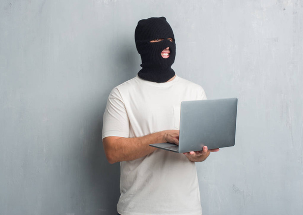 Nuori valkoihoinen hakkeri mies yli harmaa grunge seinä tekee tietoverkkohyökkäys käyttäen kannettavaa tietokonetta onnellinen kasvot pystyssä ja hymyillen luottavainen hymy osoittaa hampaat
 - Valokuva, kuva