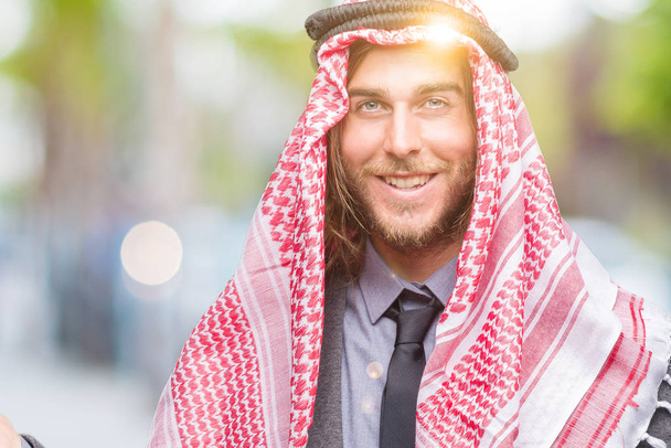 Joven hombre árabe guapo con el pelo largo usando keffiyeh sobre fondo aislado asombrado y sonriendo a la cámara mientras se presenta con la mano y señalando con el dedo
. - Foto, imagen