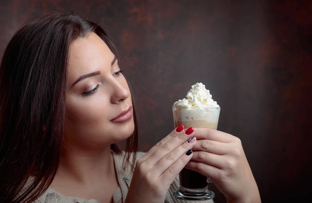 Giovane bella donna con i capelli scuri raccolti tenendo un caffè tazza con crema. Trucco perfetto, sfondo marrone
.  - Foto, immagini