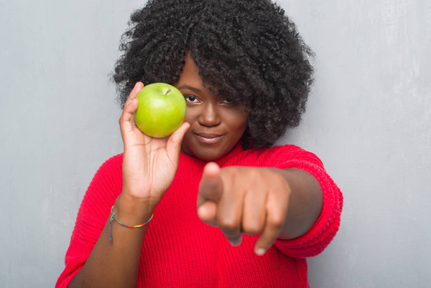 Αφρικανική αμερικανική γυναίκα πάνω από τοίχο γκρι grunge τρώει πράσινο μήλο, δείχνοντας με το δάχτυλο στη φωτογραφική μηχανή και σε εσάς, το χέρι συνδεθείτε, θετική και σίγουρη κίνηση από το μέτωπο - Φωτογραφία, εικόνα