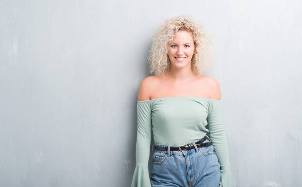 Young blonde woman with curly hair over grunge grey background with a happy and cool smile on face. Lucky person. - Photo, Image