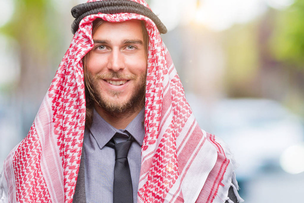 Joven hombre árabe guapo con el pelo largo usando keffiyeh sobre un fondo aislado con una sonrisa feliz y fresca en la cara. Persona afortunada
. - Foto, imagen