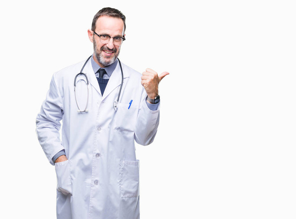 Старший седовласый врач средних лет в медицинской форме, изолированном фоне, улыбающийся со счастливым лицом, смотрящий и указывающий на сторону с большим пальцем
. - Фото, изображение