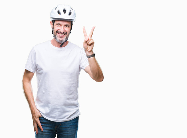 Hombre ciclista de edad madura con casco de seguridad en bicicleta fondo aislado sonriendo con la cara feliz guiñando el ojo a la cámara haciendo signo de victoria. Número dos.
. - Foto, Imagen