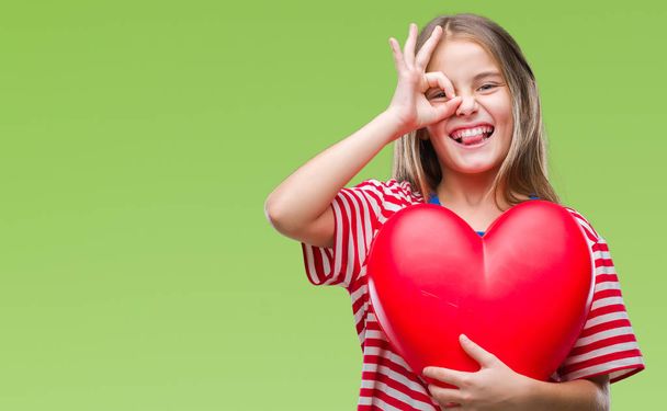 Молодая красивая девушка держит красное сердце на изолированном фоне со счастливым лицом улыбается делает ОК знак с рукой на глазу глядя через пальцы
 - Фото, изображение