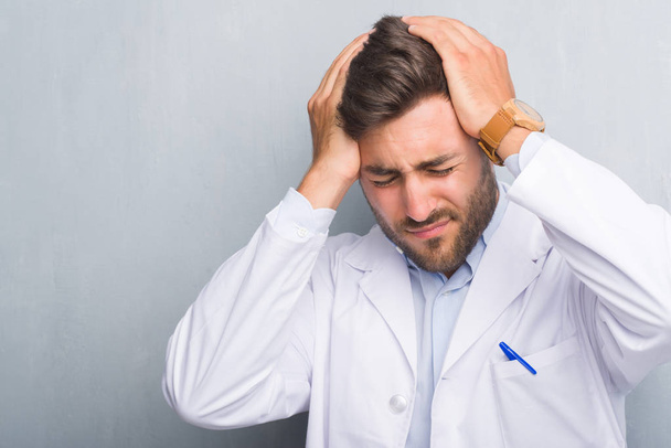 Handsome young professional man over grey grunge wall wearing white coat suffering from headache desperate and stressed because pain and migraine. Hands on head. - Photo, Image