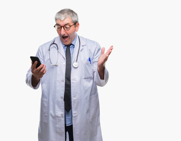 Pěkný starší lékař muž SMS odesílání zpráv pomocí smarpthone nad izolované pozadí velmi šťastný a nadšený, vítěz výraz slaví vítězství křičí s velkým úsměvem a pozvedl ruce - Fotografie, Obrázek