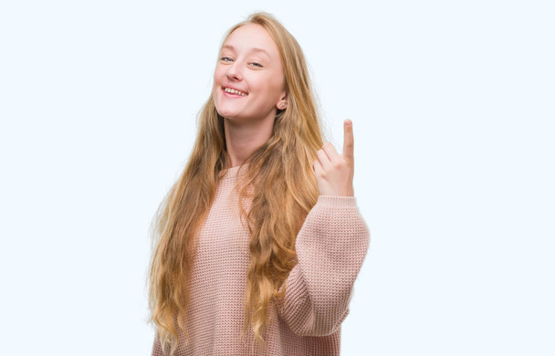 Blonde teenager woman wearing pink sweater showing and pointing up with finger number one while smiling confident and happy. - Photo, image