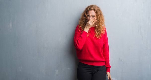 Mujer pelirroja joven sobre la pared gris grunge usando suéter rojo oliendo algo apestoso y asqueroso, olor intolerable, conteniendo la respiración con los dedos en la nariz. Concepto de malos olores
. - Foto, Imagen
