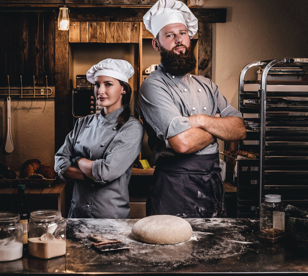Chef with his assistant in cook uniform posing with crossed arms near table with ready dough in the bakery. - Photo, Image