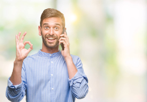 Jonge knappe man spreken op de telefoon over geïsoleerde achtergrond doen ok teken met vingers, uitstekende symbool - Foto, afbeelding