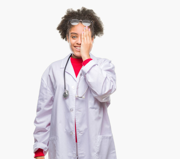 Young afro american doctor woman over isolated background covering one eye with hand with confident smile on face and surprise emotion. - Foto, afbeelding