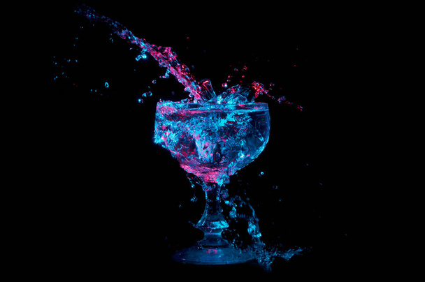 Прозора рідина під синьо-червоними вогнями розбризкується у скляний горщик ізольовано на чорному тлі
 - Фото, зображення