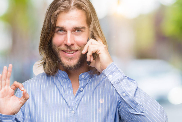 Jeune homme beau aux cheveux longs sur fond isolé parlant au téléphone faisant signe ok avec les doigts, excellent symbole
 - Photo, image