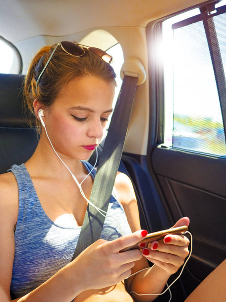 A cute teenage girl looking at the screen of her smartphone while sitting in a van wearing headphones. - Zdjęcie, obraz