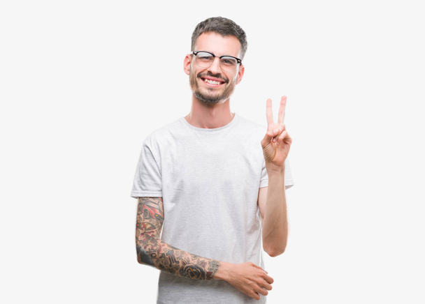Jeune homme adulte tatoué souriant avec un sourire heureux clin d'oeil à la caméra faisant signe de victoire. Numéro deux
. - Photo, image