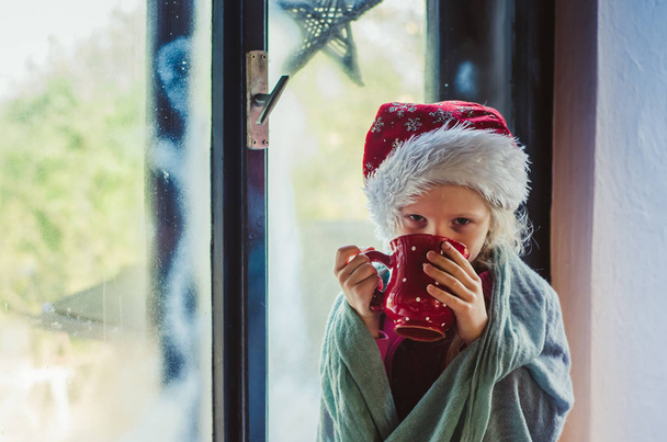 όμορφη ξανθιά κορίτσι κάθεται δίπλα στο παράθυρο με κόκκινο καπέλο Χριστουγέννων και πίνοντας ζεστό τσάι - Φωτογραφία, εικόνα
