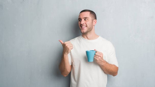 joven caucásico hombre sobre gris grunge pared driking un taza de café apuntando y mostrando con el pulgar hasta el lado con feliz cara sonriendo
 - Foto, Imagen