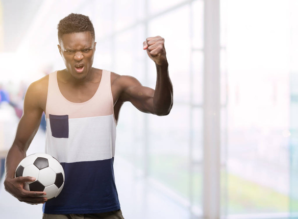Νεαρός αφρικανική αμερικανική κατοχή μπάλας ποδοσφαίρου ποδοσφαίρου ενοχλημένοι και απογοητευμένοι φωνάζει με θυμό, τρελό και φωνάζοντας με σήκωσε το χέρι, θυμό έννοια - Φωτογραφία, εικόνα