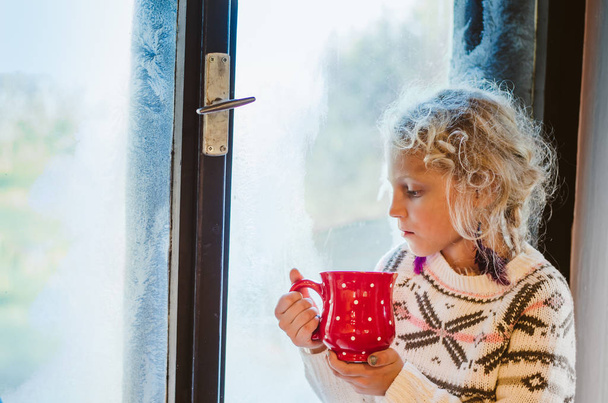 όμορφη ξανθιά κορίτσι κάθεται δίπλα στο παράθυρο και πίνοντας ζεστό τσάι από την κόκκινη διακεκομμένη κούπα - Φωτογραφία, εικόνα