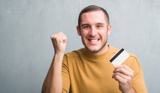 Молодий кавказький чоловік над сірою гранжевою стіною тримає кредитну картку роздратованим і розчарованим криком з гнівом, божевільним і криком з піднятою рукою, концепцією гніву
 - Фото, зображення