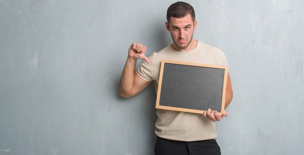 Young caucasian man over grey grunge wall holding blackboard with angry face, negative sign showing dislike with thumbs down, rejection concept - Photo, Image
