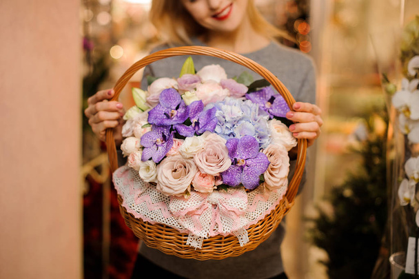 Girl in grey shirt holding in her hands a beautiful big basket of tender pink roses, violet orchids and blue hydrangeas - 写真・画像