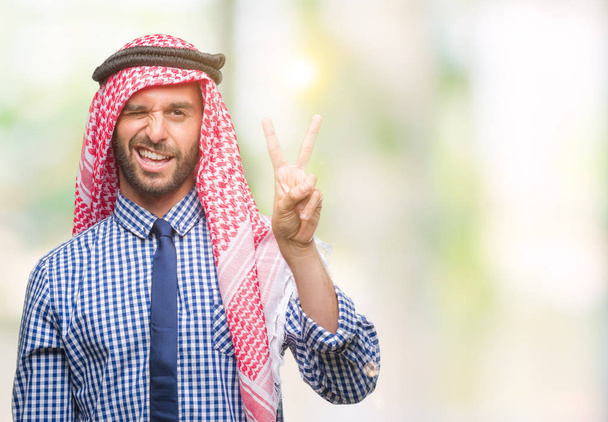 Молодой красивый арабский бизнесмен в кеффие на изолированном фоне улыбается со счастливым лицом, подмигивая в камеру, делая знак победы. Номер два:
. - Фото, изображение