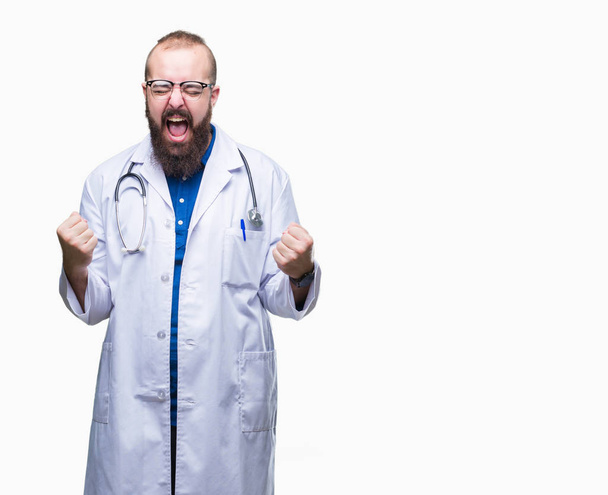 Молодий кавказький лікар у медичному білому пальто над ізольованим тлом дуже щасливий і схвильований жест переможця з піднятими руками, посміхаючись і кричачи на успіх. Концепція святкування
. - Фото, зображення
