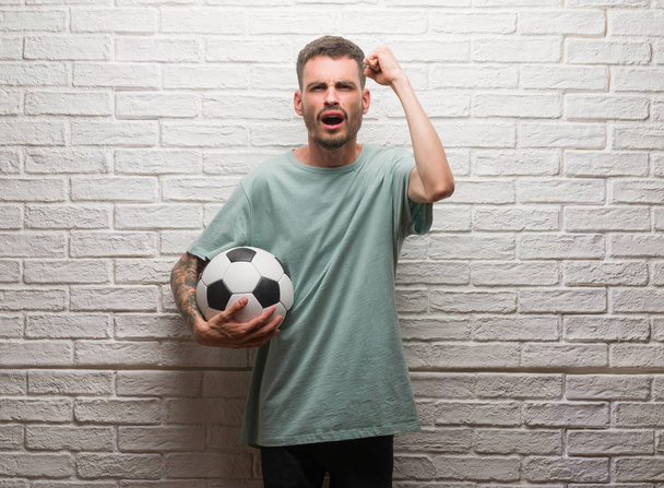 Νεαρός ενήλικος κρατώντας ποδόσφαιρο μπάλα στέκεται πάνω από το λευκό τοίχο ενοχλημένοι και απογοητευμένοι φωνάζει με θυμό, τρελό και φωνάζει με σήκωσε το χέρι, ο θυμός έννοια - Φωτογραφία, εικόνα