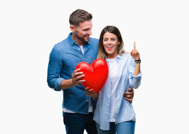 Jeune couple amoureux tenant coeur rouge sur fond isolé surpris par une idée ou une question pointant du doigt avec un visage heureux, numéro un
 - Photo, image