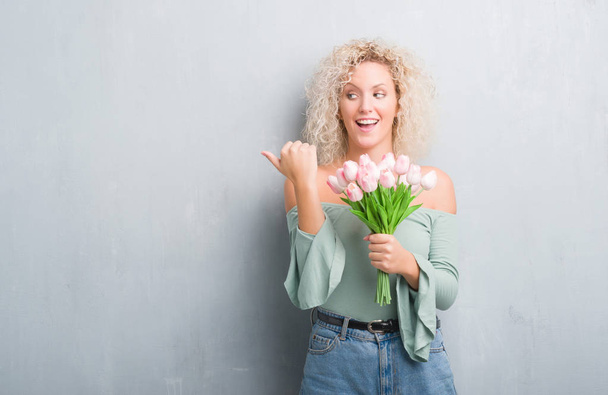 Young blonde woman over grunge grey background holding flowers pointing and showing with thumb up to the side with happy face smiling - Foto, imagen