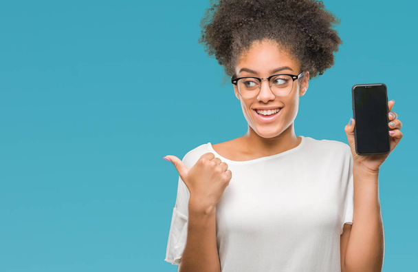 Young beautiful afro american showing smartphone screen over isolated background pointing and showing with thumb up to the side with happy face smiling - Photo, Image