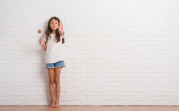 Young hispanic kid over white brick wall eating lollipop candy at home with open hand doing stop sign with serious and confident expression, defense gesture - Photo, Image