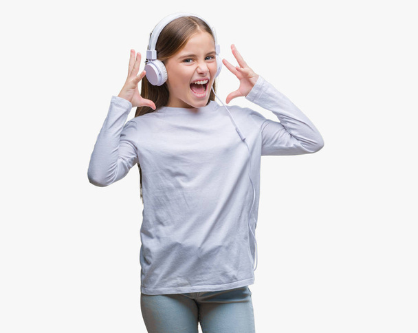 Όμορφη κοπέλα φορώντας ακουστικά να ακούτε μουσική μέσω απομονωμένη φόντο γιορτάζει τρελών και τρελό για την επιτυχία με τα όπλα έθεσε και έκλεισε τα μάτια ουρλιάζοντας ενθουσιασμένος. Νικητής έννοια - Φωτογραφία, εικόνα