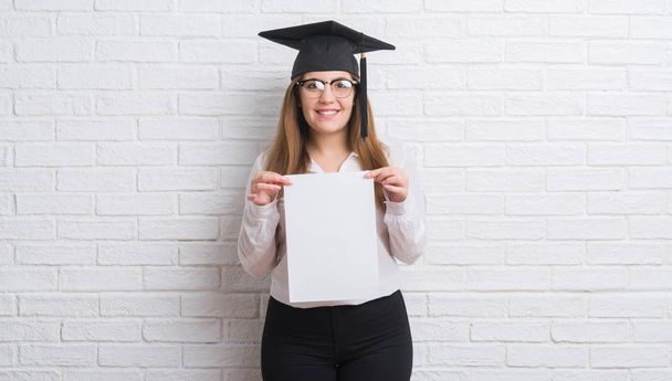 Fiatal felnőtt nő visel végzős kap gazdaság diploma, boldog arccal állt, és mosolyogva, magabiztos mosollyal mutatja a fogak fehér tégla falon - Fotó, kép