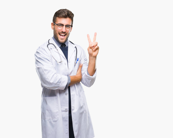 Young handsome doctor man over isolated background smiling with happy face winking at the camera doing victory sign. Number two. - Фото, изображение