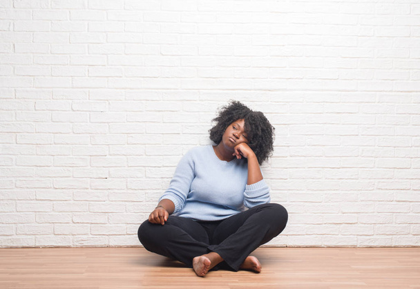 Νεαρός αφρικανική αμερικανική γυναίκα που κάθεται στο πάτωμα στο σπίτι νομίζοντας ψάχνει κουρασμένη και τρυπημένο με προβλήματα κατάθλιψης με σταυρωμένα χέρια. - Φωτογραφία, εικόνα