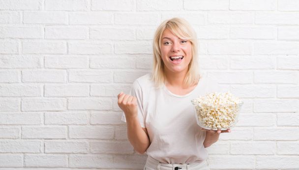 Adult caucasian woman over white brick wall eating pop corn screaming proud and celebrating victory and success very excited, cheering emotion - Фото, зображення