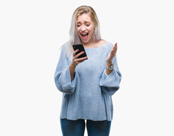 Mladá blond žena SMS odesílání zpráv pomocí smartphone přes samostatný pozadí velmi šťastný a nadšený, vítěz výraz slaví vítězství křičet s velkým úsměvem a pozvedl ruce - Fotografie, Obrázek