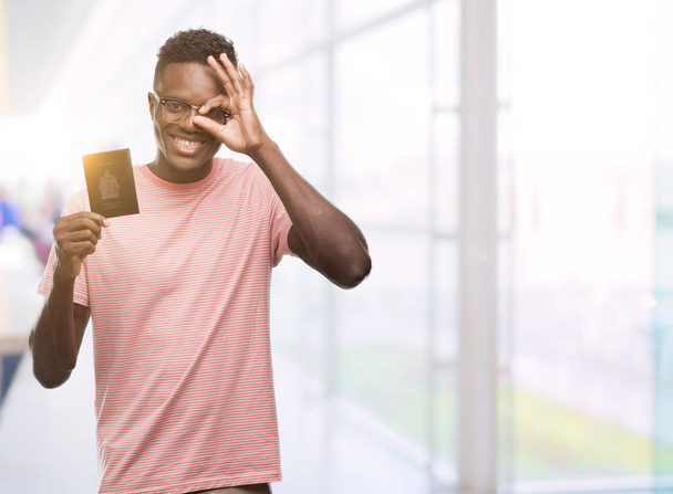 Young african american man holding canadian passport with happy face smiling doing ok sign with hand on eye looking through fingers - Photo, Image
