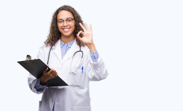 Joven doctora hispana sosteniendo un portapapeles haciendo buen signo con los dedos, excelente símbolo
 - Foto, imagen