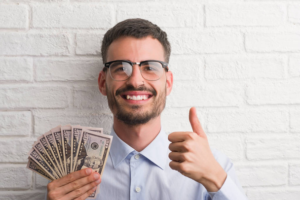 Giovane uomo d'affari hipster in possesso di dollari felice con grande sorriso facendo segno ok, pollice in su con le dita, segno eccellente
 - Foto, immagini