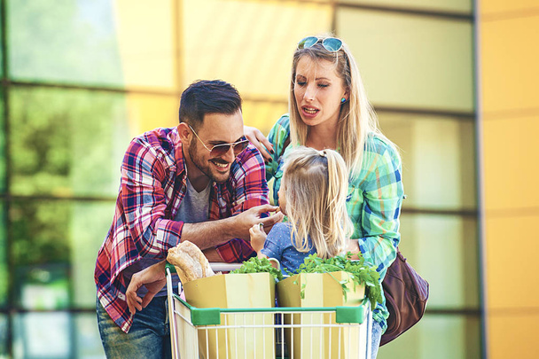Young smiling parents and their cute daughter with shopping cart full of fresh food having fun in front of hypermarket. - 写真・画像