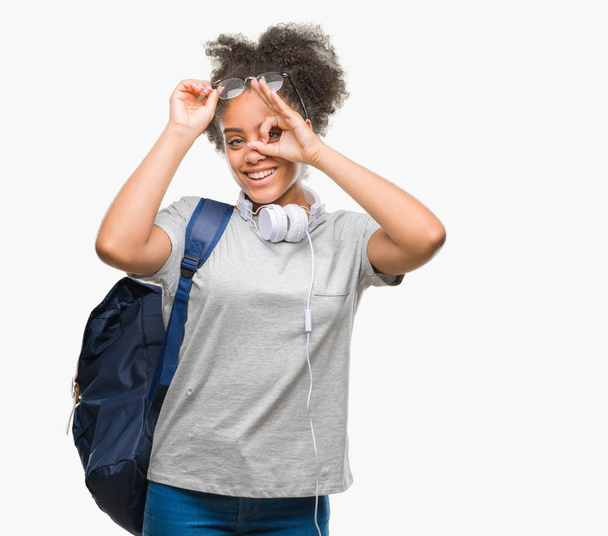 Young afro american student woman wearing headphones and backpack over isolated background doing ok gesture with hand smiling, eye looking through fingers with happy face. - Photo, Image