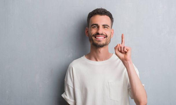 Mladý dospělý muž stojící nad zeď šedá grunge překvapilo s nápad nebo otázku ukazující prst s šťastné tváře, číslo jedna - Fotografie, Obrázek