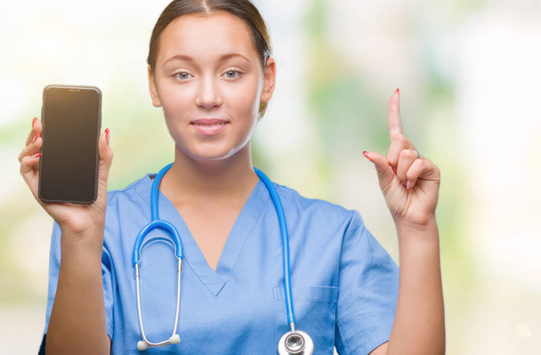 Jovem mulher médica caucasiana mostrando smarpthone sobre fundo isolado surpreendido com uma ideia ou pergunta apontando o dedo com o rosto feliz, número um
 - Foto, Imagem