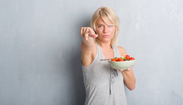 erwachsene kaukasische Frau über grunge-grauer Wand, die Tomatensalat isst und mit dem Finger in die Kamera und zu dir zeigt, Handzeichen, positive und selbstbewusste Geste von vorne - Foto, Bild